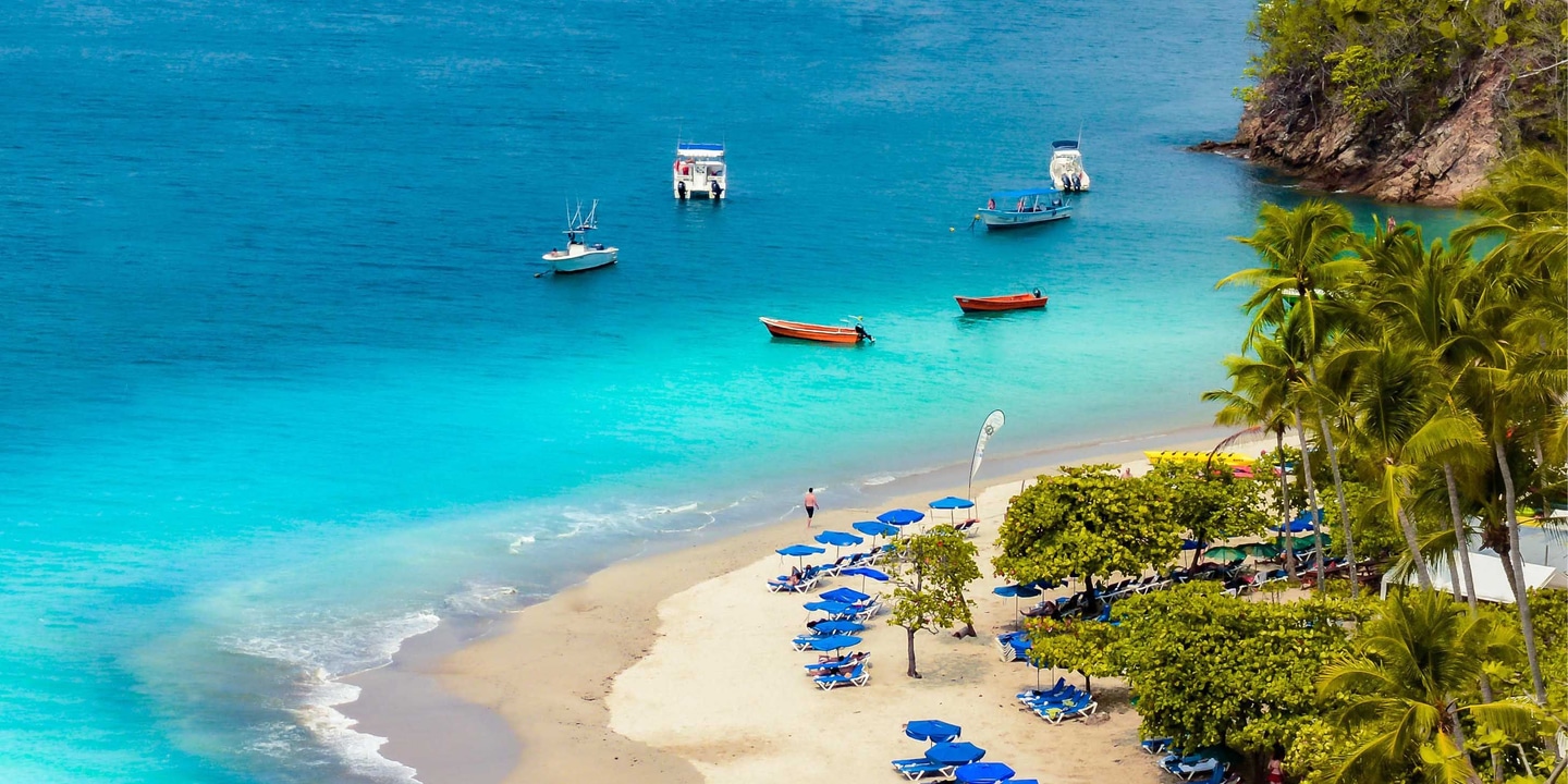 Small fishing boats in the crystal-clear blue waters and sunbeds on a palm tree lined white sandy beach in Tortuga in the Caribbean
