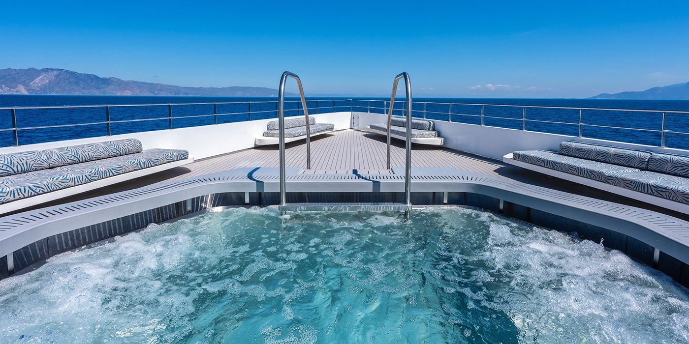 Observation deck pool on board Emerald Kaia, with bubbling water, cushioned lounge seating and panoramic ocean and coastline views under a clear blue sky.