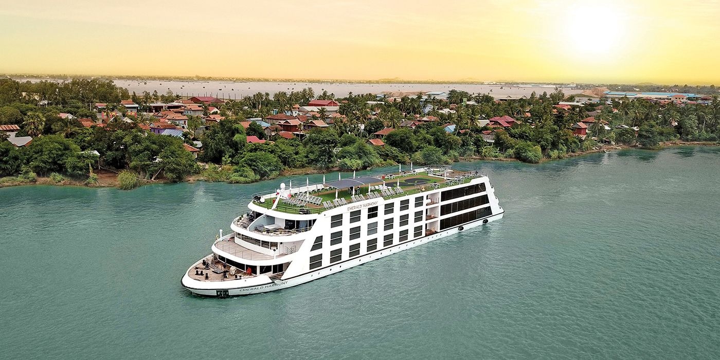 Emerald Harmony river cruise ship gliding along a calm, teal-green Mekong River at sunset, passing a lush riverside village of palm trees and colourful houses under a golden sky.