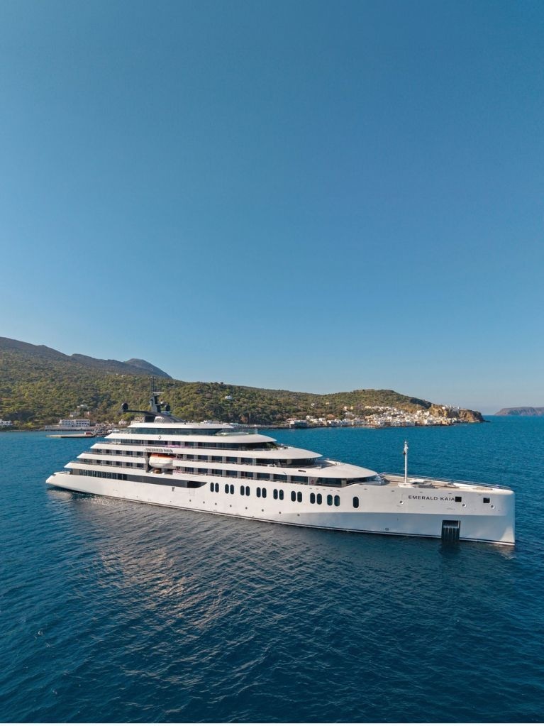 A modern luxury cruise ship anchored in calm blue waters near a sunlit coastal landscape.